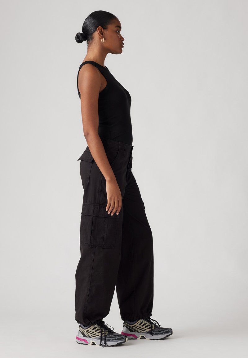 Un débardeur noir à côtes associé à un pantalon cargo noir ample avec des poches latérales, et des baskets grises avec des accents roses. Vue de côté complète.