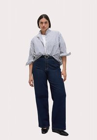 Stripet skjorte med knapper og op rullede ærmer, kombineret med højtsiddende mørkeblå brede bukser og sorte sko. Simpelt design og neutrale farver.