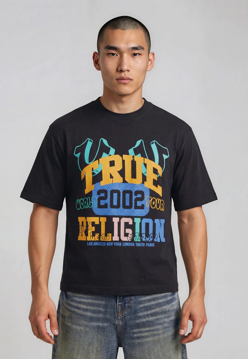Uomo con capelli corti che indossa una t-shirt nera con grafica True Religion e jeans blu sbiaditi, in piedi davanti a uno sfondo grigio uniforme.