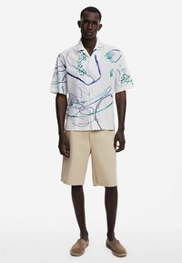 Kortærmet hvid skjorte med knapper, dekoreret med grønne og blå abstrakte mønstre, kombineret med beige shorts og lysebrune slip-on sko.