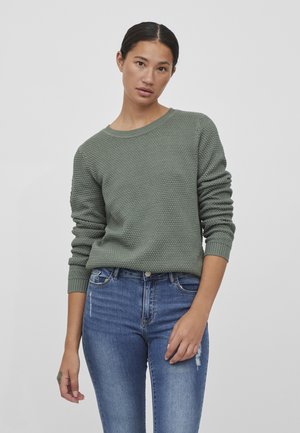 Pulover texturat verde cu guler rotund și manșete dantelate, purtat cu blugi din denim albastru cu ușoare deteriorări.