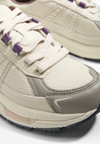 Krémová a šedá běžecká obuv z materiálů síťoviny a kůže, s fialovými akcenty, texturovanými povrchy a silnou gumovou podrážkou.