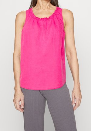 Femme portant un haut sans manches rose vif avec un col froncé et un pantalon gris côtelé, debout devant un fond neutre.