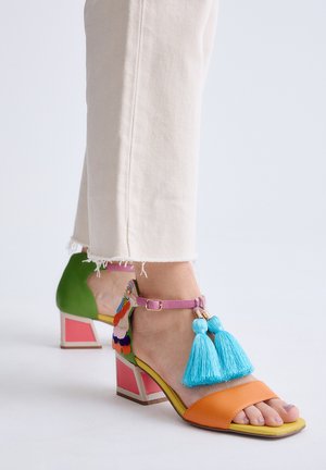 Pie con sandalia de tacón de bloque colorido con tira naranja en los dedos, tira rosa en el tobillo, borlas azules y detalle multicolor en el tacón, sobre pantalones blancos.