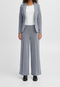 Blazer gris en maille avec pantalon large assorti. Les deux présentent une texture lisse, un design structuré et des poches latérales. Associés à un haut blanc.