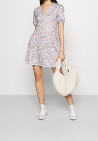 Blommig klänning med V-ringning, korta puffärmar och utsvängd kjol, med rosa och gröna mönster på en ljusblå bakgrund. Plyschväska i krämfärg.