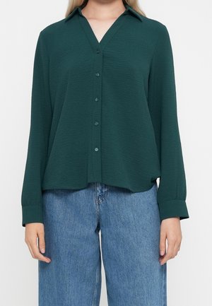 Femme portant une chemise texturée à manches longues de couleur vert foncé avec col et jean en denim bleu, debout devant un fond uni.