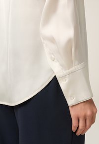 Blusa di seta bianca con orlo rotondo, polsini abbottonati con dettagli di cucitura argentata, abbinata a pantaloni blu scuro.