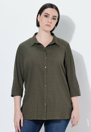 Young woman with dark hair wears an olive green textured button-up shirt with 3/4 sleeves and blue jeans, standing against a white wall.