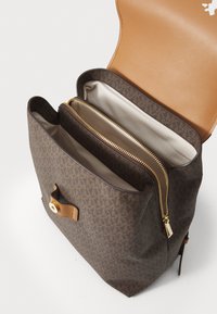 Sac à dos marron à motif logo avec un rabat en cuir tan, comportant un intérieur zippé et plusieurs compartiments. Accents en métal doré visibles.