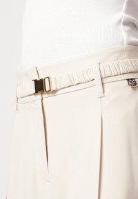 Beige Hosen mit einem gerafften Taillenbund und goldenen Metallverschlussdetails. Sie verfügen über Seitentaschen und eine glatte Stofftextur.
