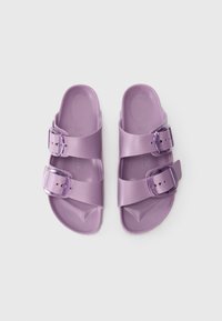Violets atvērta pirksta sandales ar divām regulējamām sprādzēm, konturēta zole un teksturēts pamatne, rādītas no augšas uz balta fona.