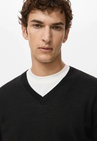 Jonge man met krullend bruin haar, gekleed in een zwarte V-hals trui over een wit shirt met ronde hals, tegen een effen lichte achtergrond.