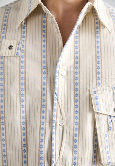 Camicia beige con colletto, decorata da motivi floreali blu disposti lungo strisce verticali, con tasca frontale e bottoni bianchi.