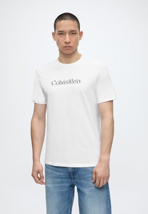Mann, der nach vorne schaut, trägt ein weißes Calvin Klein T-Shirt und hellblaue Jeans vor einem schlichten Hintergrund.
