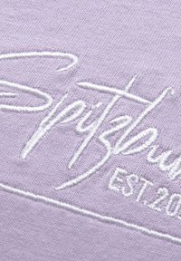 Lavender cotton fabric features white embroidered text "Spitfire" and "EST. 2000," with a smooth texture and subtle stitch detail.