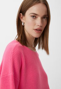 Rosa gestrickter Pullover mit weitem Ausschnitt und strukturiertem Stoff. Das Model hat schulterlanges braunes Haar und schlichte silberne Creolen.