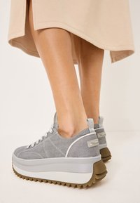 Grå canvas sneakers med en tjock vit mellansula och sågad brun yttersula. Har vita snören och en dragflik på hälen.