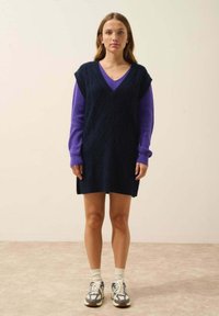 Granatowa dzianinowa sukienka bez rękawów z dekoltem w szpic, z diamentowym wzorem. Fioletowa bluzka z długim rękawem pod spodem. Jasne trampki.