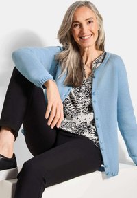 Gros cardigan en maille bleu clair avec un motif texturé, fermeture à boutons, porté sur une blouse à motif monochrome et un pantalon noir. Tissu doux.