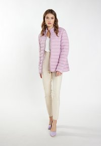 Lys lilla pufferjakke med høj krave, lynlås foran og vandret quiltet mønster, parret med beige slim-bukser og lavendel hæle.