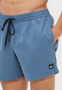 Lyseblå badebukser laget av glatt stoff, med elastisk midjebånd og svarte snorer, samt et lappet logo på venstre bein.