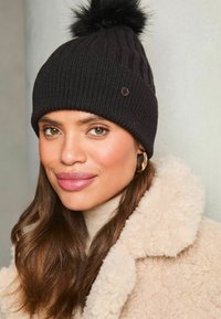 Gorro de punto negro con canalé vertical, rematado con un pompón esponjoso y un acento de logo de marca metálico. Textura de lana, diseño ajustado.