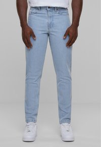 Jeans in denim azzurro chiaro con vestibilità slim, dotati di cinque tasche, chiusura con bottone e zip, e dettagli cuciti. Indossati con sneakers bianche.