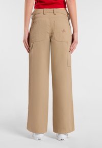 Beige cargo broek met wijde pijpen, patchzakken, zichtbare stiksels en een logo patch op de rechterzak. Gladde textuur, casual ontwerp.