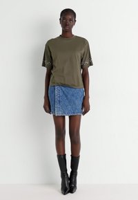Olijfgroene t-shirt met studs op de mouwen, gecombineerd met een jeansrok met een blauw ruitpatroon en een zijrits, gedragen met zwarte laarzen.
