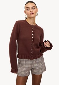 Cardigan marron en maille avec ourlet et poignets à volants, doté de boutons en perle sur le devant. Associé à un short à carreaux.