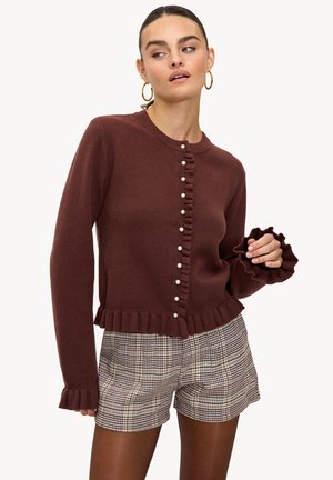 Cardigan marron en maille avec ourlet et poignets à volants, doté de boutons en perle sur le devant. Associé à un short à carreaux.