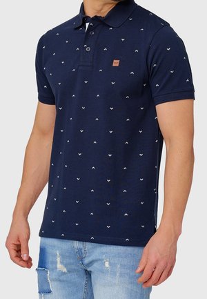 Mand iført marineblå poloshirt med små hvide chevronmønstre og lyseblå revnede jeans mod ensartet baggrund.