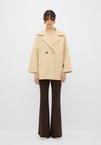 Cappotto doppiopetto beige con colletto oversize, grandi tasche e maniche larghe, abbinato a pantaloni brown a zampa d'elefante e tacchi bianchi.