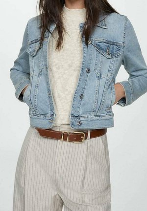 Person iført en lyseblå denimjakke over en cremefarvet sweater, beige bukser med striber og brunt bælte med guldlås, hænder i jakkelommerne.