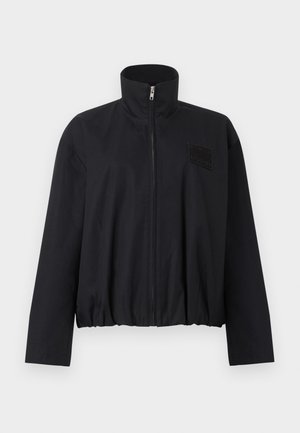 Must musta lukuga jakk kõrge kraega, pikkade varrukatega, elastse äärega ja vasakul rinnal väikese plaastriga.