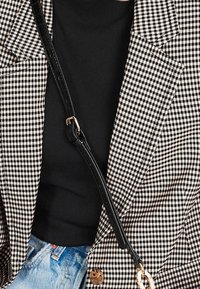 Haut noir à col montant sous un blazer en pied-de-poule noir et blanc avec une sangle noire traversant le corps et un jean denim bleu clair visible à la taille.