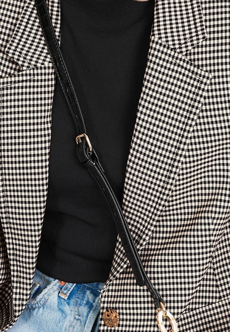 Haut noir à col montant sous un blazer en pied-de-poule noir et blanc avec une sangle noire traversant le corps et un jean denim bleu clair visible à la taille.