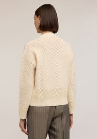 Pletený cardigan v krémové barvě s otevřeným síťovaným vzorem, s ribovaným lemem a manžetami. Stylizováno s proužkovanými kalhotami, pohled zezadu.