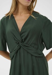 Rochie verde fabricată dintr-un material neted, cu un decolteu adânc în V, mâneci scurte și un detaliu cu talie legată, care oferă o croială lejeră.