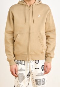 Beżowa bluza z kapturem na zamek, z kieszenią kangurzą, kapturem ze sznurkiem i białym logo na piersi. Miękka, gładka faktura; luźny krój.