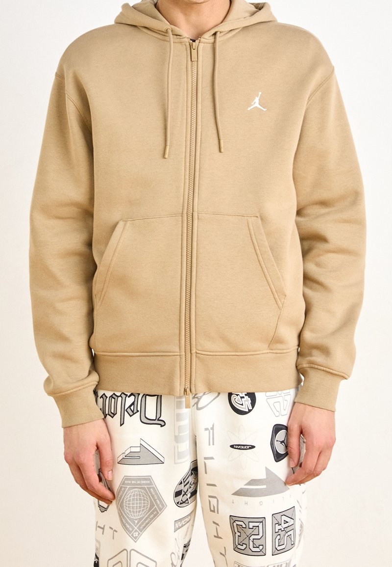 Beżowa bluza z kapturem na zamek, z kieszenią kangurzą, kapturem ze sznurkiem i białym logo na piersi. Miękka, gładka faktura; luźny krój.