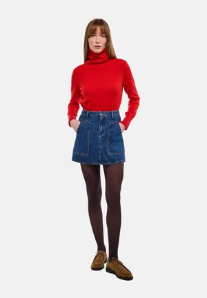 Femme debout, mains dans les poches, portant un pull à col roulé rouge, une jupe en denim bleu, des collants noirs et des chaussures marron.