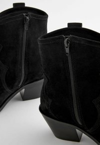 Botas de tobillo de ante negro con una textura suave, cremalleras laterales y un tacón apilado. Presentan detalles de costura y un diseño de punta redonda.