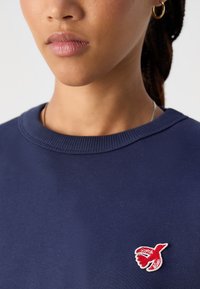 Σκούρο μπλε φούτερ με ριχτό λαιμό και μικρό κόκκινο κεντητό λογότυπο ψαριού στο αριστερό στήθος. Λείο υλικό.