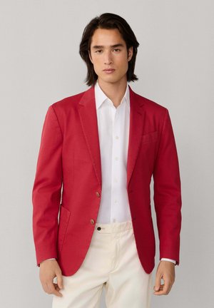 Jeune homme aux cheveux noirs mi-longs portant un blazer rouge, une chemise blanche et un pantalon crème, debout devant un fond uni.