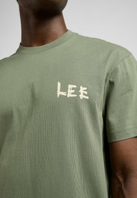 Oliwkowa bawełniana koszulka z okrągłym dekoltem. Posiada jasnobeżowy nadrukowany logotyp "LEE" na lewej stronie klatki piersiowej. Miękka tekstura, luźny krój.