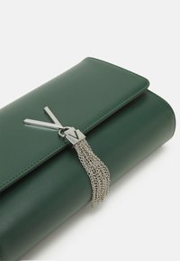 Zelená kožená kabelka na ruku s stříbrným řetízkovým třásněním a výrazným stříbrným akcentem ve tvaru 'V'. Hladký povrch s přehybovým uzávěrem.
