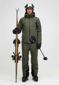 Ski-outfit in olijfgroen, met een waterdichte jas en broek. Houdt zwarte skistokken en ski's met oranje accenten en bindingen vast.
