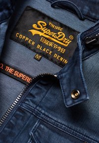 Inredning av denimjacka med en svart etikett med "SuperDry" i gult, storlek "M," och koppardetaljer på mörkblått tyg. Dragkedja synlig.
